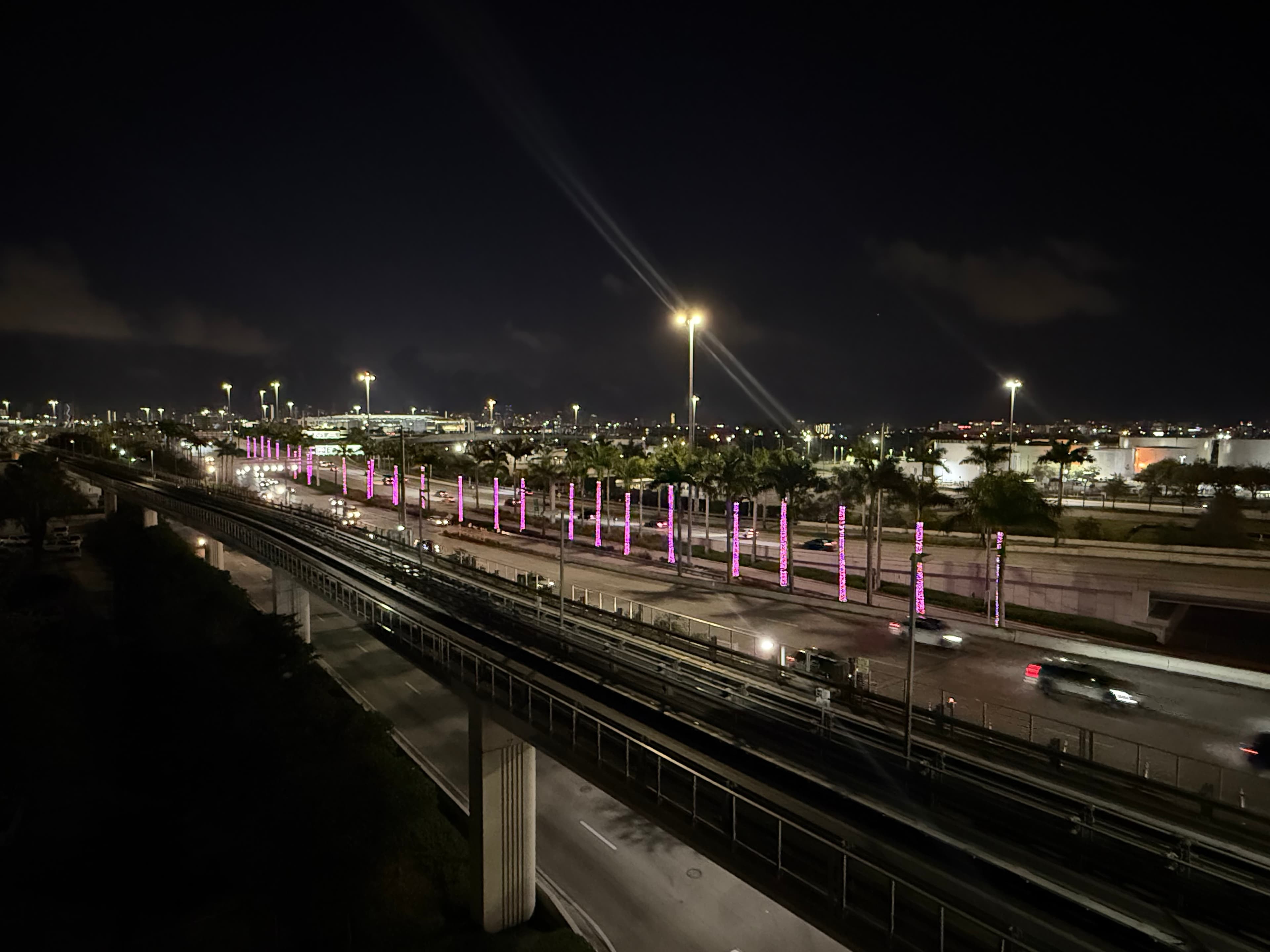 Nighttime view of elevated train tracks above a highway lined with pink-lit palm trees.