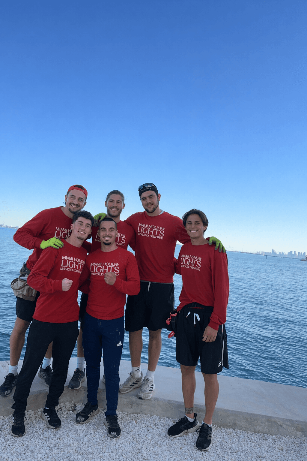 Six men in red Miami Holiday Lights shirts pose by the ocean with city skyline.